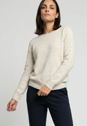 Vrouw draagt een lichtbeige geribde trui en donkere broek, staand tegen een effen lichte achtergrond, poserend met één hand bij haar taille.