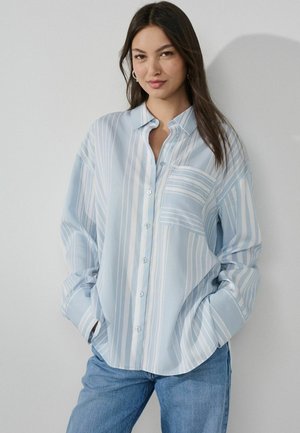 Young woman with dark hair wears light blue and white striped button-up shirt and blue jeans, standing with hands in pockets against gray wall.