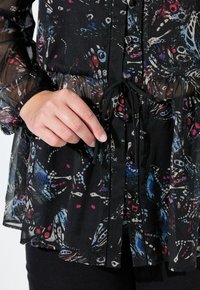 Černá blůza s květinovými a abstraktními vzory v modré, červené a bílé barvě. Má stažený pas, zapínání na knoflíky a průsvitné rukávy.