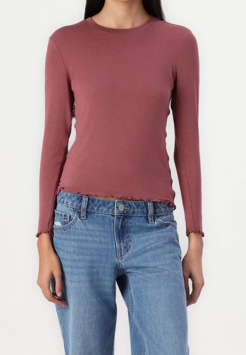 Långärmad topp i rostfärg av mjuk bomull, med rund hals och volang nertill, tillsammans med ljusblå jeans med hög midja.