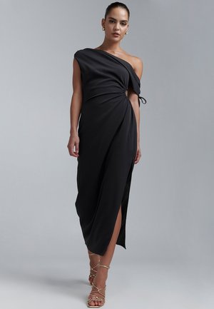 Črna obleka z enim naramnicami, z razpeto tkanino, stranskim razporkom in detajlom vozla v predelu pasu. Oblečena s sandali z zlatimi trakovi.