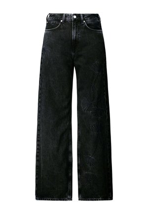 Personne portant un jean noir à jambes larges avec une broderie florale subtile sur la cuisse, une blouse blanche rentrée, et une ceinture noire, main dans la poche.
