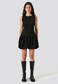 Abito nero senza maniche con corsetto aderente e gonna a volant. Abbinato a stivali neri in pelle al ginocchio. Texture liscia e design semplice.