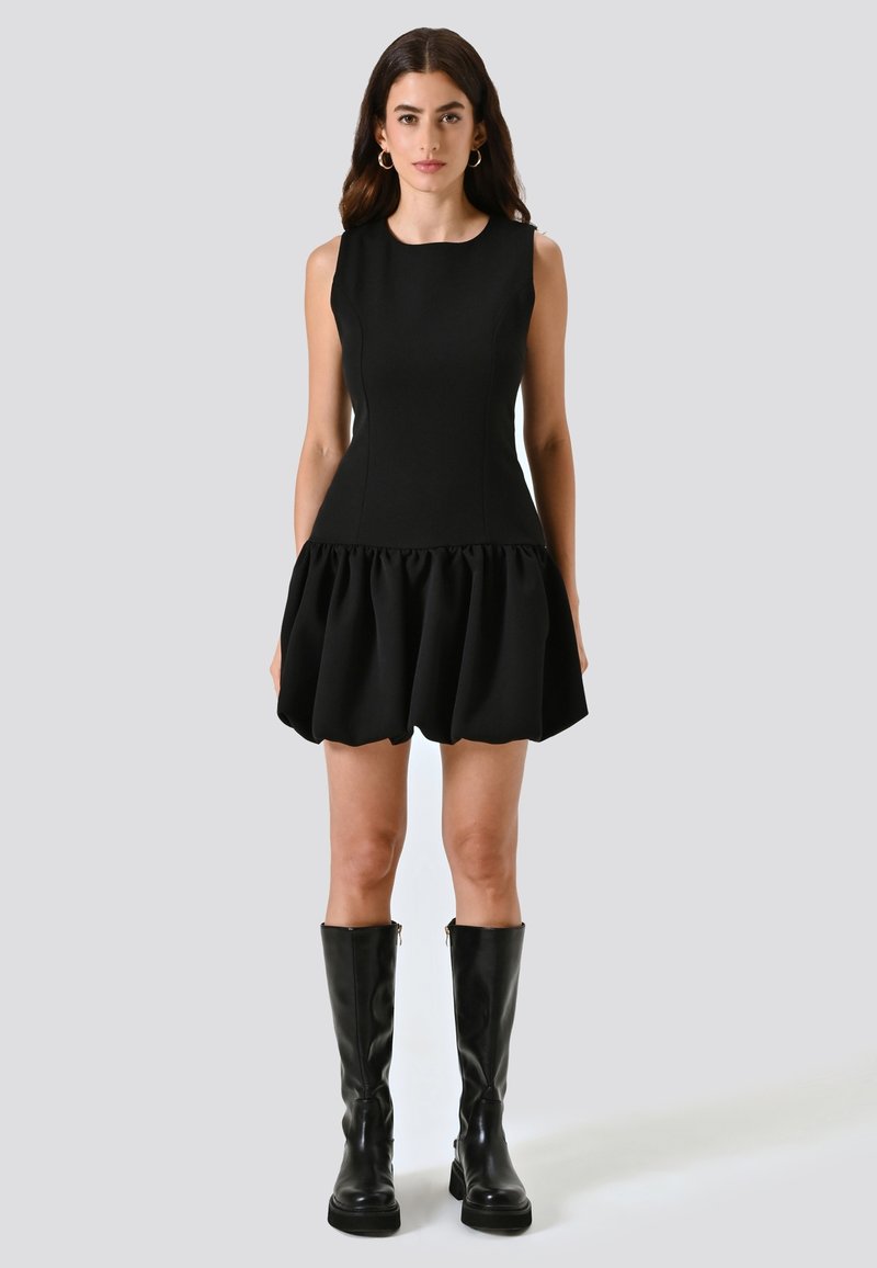 Abito nero senza maniche con corsetto aderente e gonna a volant. Abbinato a stivali neri in pelle al ginocchio. Texture liscia e design semplice.