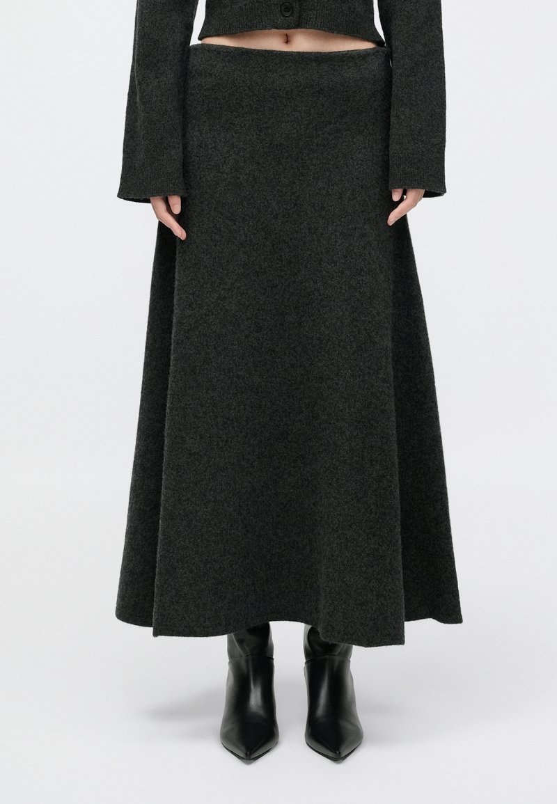 Σκούρο γκρι, μακριά πλεκτή φούστα με φαρδύ ζωνάκι και ροή σιλουέτας, συμπληρωμένη από μυτερές μαύρες μπότες μέχρι τον αστράγαλο.