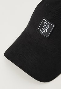 Black corduroy cap featuring a square patch with a white logo. The bill has a slight curve with textured fabric.