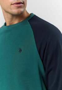 Zaļš un jūras zila kokvilnas crewneck džemperis, ar ribotu apkakli un izšūtu logo uz krūtīm, gluda tekstūra, ikdienišķs dizains.