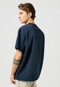 Camiseta de algodón azul marino, corte relajado, mangas cortas, escote redondo, con costuras visibles y un dobladillo plano, llevada con pantalones de color claro.