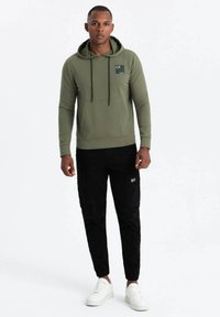 Felpa verde oliva con tasca frontale e cordini neri, abbinata a pantaloni cargo neri e sneaker bianche.