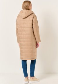 Cappotto puffa lungo beige con cappuccio, caratterizzato da una texture trapuntata orizzontale e fessure laterali. Abbinato a jeans blu e sneakers chiare.