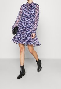 En halvgenomskinlig klänning med ett lila leopardmönster, med långa ärmar och en utställd nederdel, kombinerad med svarta ankelskor och en svart handväska.