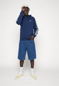 Mörkblå hoodie med vit tre-stripsdetalj, ihopparad med denimshorts och gula sneakers. Modellen bär vita strumpor och en ljus bucket hat.