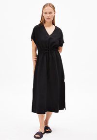Schwarzes mittelanges Kleid mit V-Ausschnitt, kurzen Ärmeln, Kordelzug in der Taille und seitlichem Schlitz. Getragen mit schwarzen Slides. Einfacher, leichter Stoff.