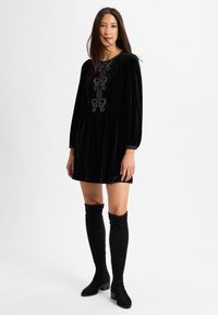 Schwarzes Samtkleid mit langen Puffärmeln, das drei dekorative Schleifen an der Vorderseite und einen plissierten Rock hat. Getragen mit schwarzen Overknee-Stiefeln.