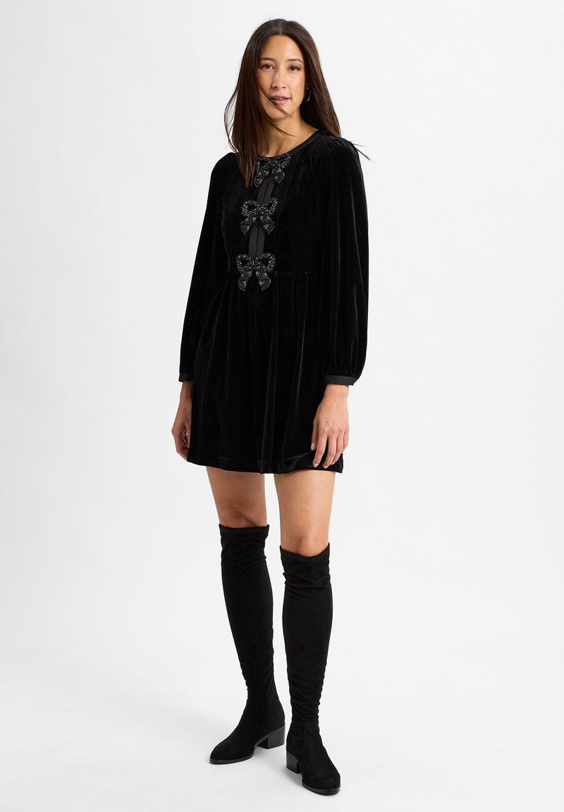 Schwarzes Samtkleid mit langen Puffärmeln, das drei dekorative Schleifen an der Vorderseite und einen plissierten Rock hat. Getragen mit schwarzen Overknee-Stiefeln.