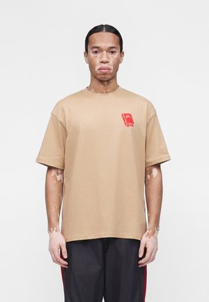 Homme atteint de vitiligo portant un t-shirt beige avec un motif rouge et un pantalon noir, debout devant un fond blanc uni.