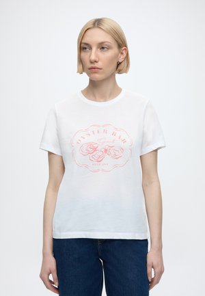 Blondynka w białym T-shircie z krótkim rękawem z różową grafiką ostrygi i tekstem, w połączeniu z ciemnoniebieskimi jeansami, stojąca na białym tle.