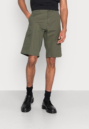 Shorts cargo vert olive avec poches latérales, coupe décontractée et passants pour ceinture. Portés avec des chaussettes noires et des chaussures noires à lacets.
