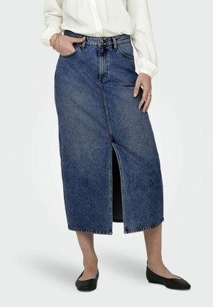 Femme portant une jupe en jean bleue mi-longue avec une fente devant, un chemisier blanc et des chaussures plates noires, debout avec une main dans la poche.