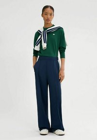Suéter verde, pantalones de pierna ancha azul marino y zapatillas blancas. El suéter está acompañado de una bufanda a rayas azul y blanca que cae sobre los hombros.
