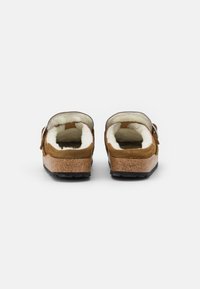 Zoccoli in suede marrone con suola in sughero, caratterizzati da un morbido rivestimento interno bianco e una fascia regolabile. Suola testurizzata per una migliore aderenza.
