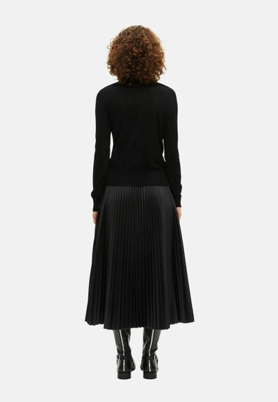 Maglione nero a maglia abbinato a una gonna nera plissettata, con pieghe orizzontali e texture lucida. Stivali neri al ginocchio con fibbie.