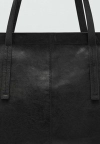 Sac fourre-tout en cuir noir avec une texture lisse, deux longues poignées et un design minimaliste. Présente des coutures visibles le long des bords.