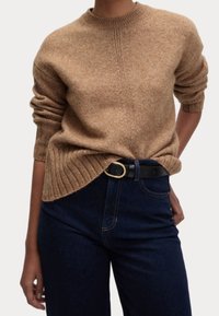 Brązowy sweter dzianinowy z okrągłym dekoltem i podwiniętymi rękawami, w połączeniu z ciemnoniebieskimi dżinsami i czarnym paskiem z złotą klamrą.