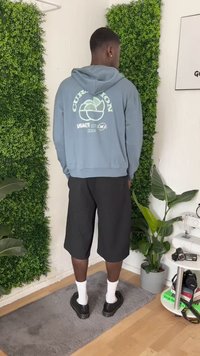 Hombre vestido con una sudadera azul claro con texto gráfico, pantalones cortos negros, calcetines blancos y zapatos negros, de pie sobre una alfombra gris en una habitación con plantas.
