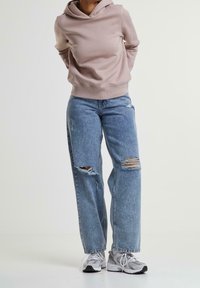 Sudadera beige con un bolsillo tipo canguro en la parte frontal, combinada con jeans rasgados de color azul claro. Zapatillas con una mezcla de tonos grises y blancos completan el look.