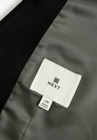 Czarna tkanina z satynowym wykończeniem oraz biała metka z napisem "NEXT" i rozmiarem "40R", wskazująca na markę i specyfikację rozmiaru produktu.