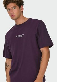 Lilla bomulds T-shirt med korte ærmer og hvid broderet tekst "CURATED STAPLES" på brystet. Afslappet pasform, blød tekstur.