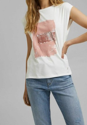 T-shirt bianco a maniche corte con un design rettangolare testurizzato in rosa e grigio con testo. Indossato con jeans di denim azzurri.