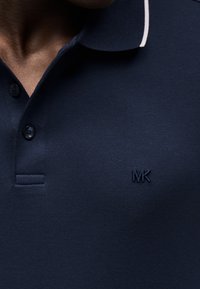 Gros plan d'une personne portant un polo bleu marine avec des boutons noirs et un petit logo brodé "MK" sur la poitrine.