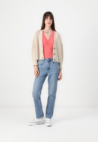 Beige strikket cardigan med ballongsleeves, parret med en koralfarvet top og lyseblå jeans, komplementeret af hvide sneakers.