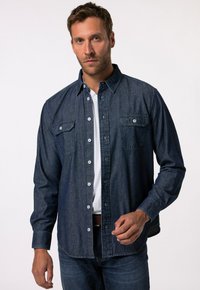 Camisa de mezclilla de botones en azul oscuro con manga larga, dos bolsillos en el pecho y botones blancos en contraste. Usada sobre una camiseta blanca.