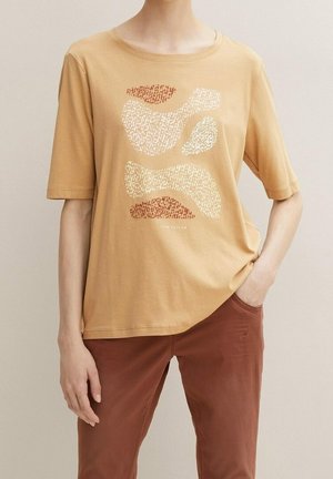 Persona que lleva una camisa beige de manga corta con formas abstractas en crema y marrón, combinada con pantalones de color óxido, con la mano en el bolsillo.