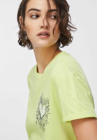 Limonkowo-zielona bawełniana koszulka z krótkimi rękawami, z nadrukiem serca z błyszczącymi akcentami. Okrągły dekolt i luźny krój.
