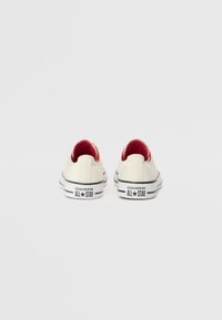 Tejszínes vászon felsőrésszel rendelkező sneaker, piros belső béléssel, fekete-fehér gumitalppal és logóval a saroknál. Kerek orrú és alacsony szárú dizájn.