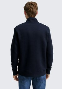 Sudadera texturizada de color azul marino con un cuello alto, puños acanalados y un acabado de tela suave. Presenta un pequeño parche de logo en la manga.