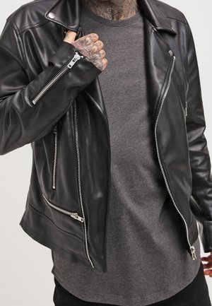 Hombre con una chaqueta de cuero negra de motociclista sobre una camiseta gris de manga larga, ajustando con la mano el cuello de la chaqueta, con tatuajes visibles en el cuello y la mano.