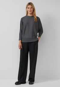 Grijze gebreide trui met lange mouwen, ronde hals en geribde zoom, gecombineerd met zwarte brede broek en zwarte schoenen, voor een effen achtergrond.
