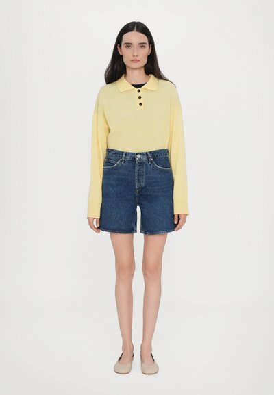 Polo gialla a maniche lunghe con bottoni, abbinata a pantaloni corti in denim blu. Il modello indossa delle ballerine beige. Design semplice e look informale. Sfondo neutro.