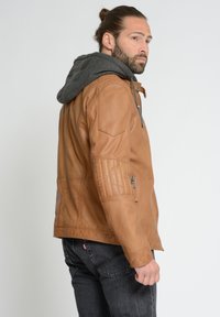 Braune Lederjacke mit grauer Kapuze, ausgestattet mit einem Reißverschluss und gesteppte Schulterdetails. Das Model trägt dunkle Jeans und zeigt ein Profil.