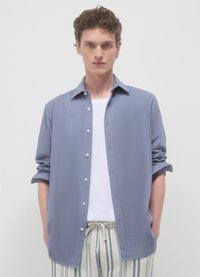 Chemise bleu clair à boutons avec un design décontracté, manches retroussées et tissu texturé, portée par-dessus un débardeur blanc et un pantalon à rayures.