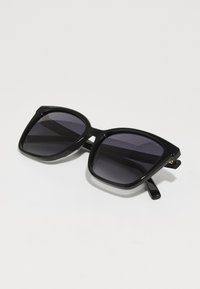 Lunettes de soleil noires surdimensionnées avec des verres carrés, teintées foncées, présentant une finition brillante et un design de monture épaisse. Branches fines et droites.