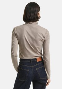 Person mit schulterlangen dunklen Haaren, der einen taupefarbenen Langarmrollkragenpullover und dunkelblaue Jeans mit einem braunen Leder-Brandpatch trägt.