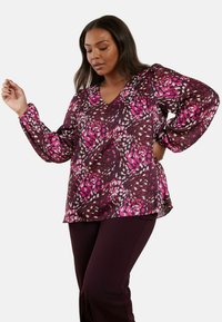 Blusa a maniche lunghe, con scollo a V, di colore viola scuro con un motivo floreale in rosa, bianco e crema. Tessuto leggero con dettagli delle maniche arricciate.