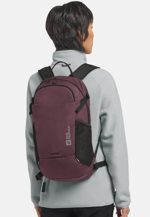 Person wearing a maroon Jack Wolfskin backpack with black mesh side pockets and padded straps, dressed in a light gray fleece jacket.