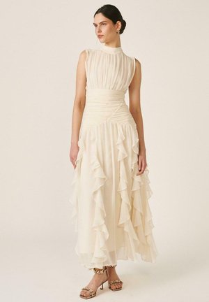 Mujer con vestido sin mangas color marfil con corpiño fruncido y falda con volantes en capas, de pie frente a un fondo liso color beige.